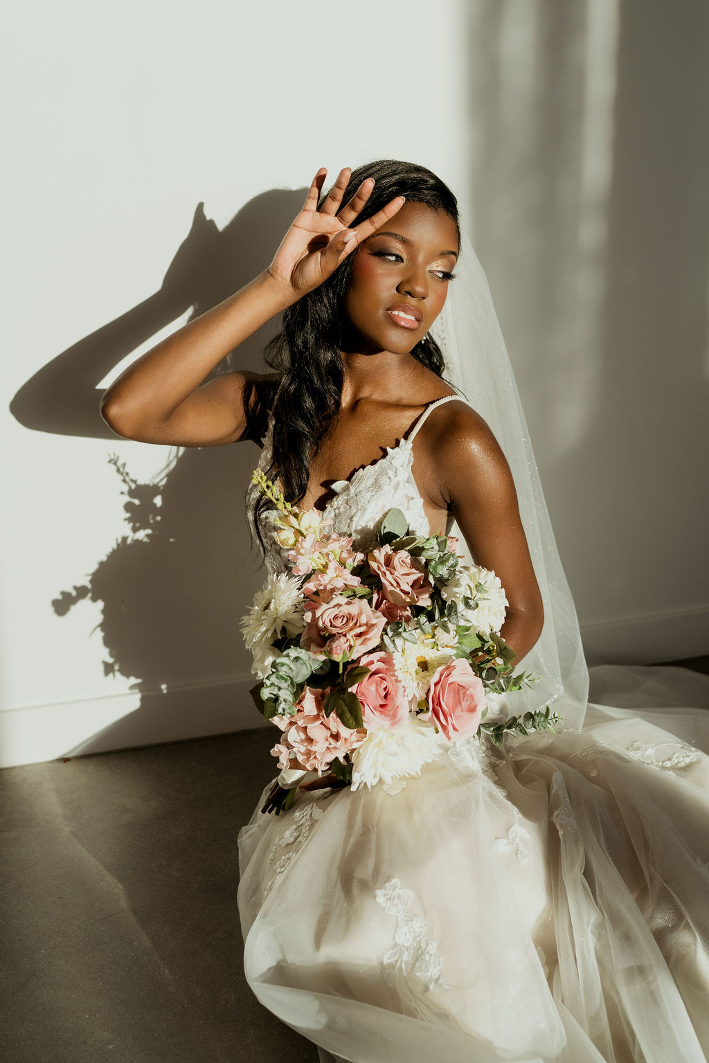 Bride in a flowing gown and veil holding a colorful bouquet in a sunlit studio portrait.